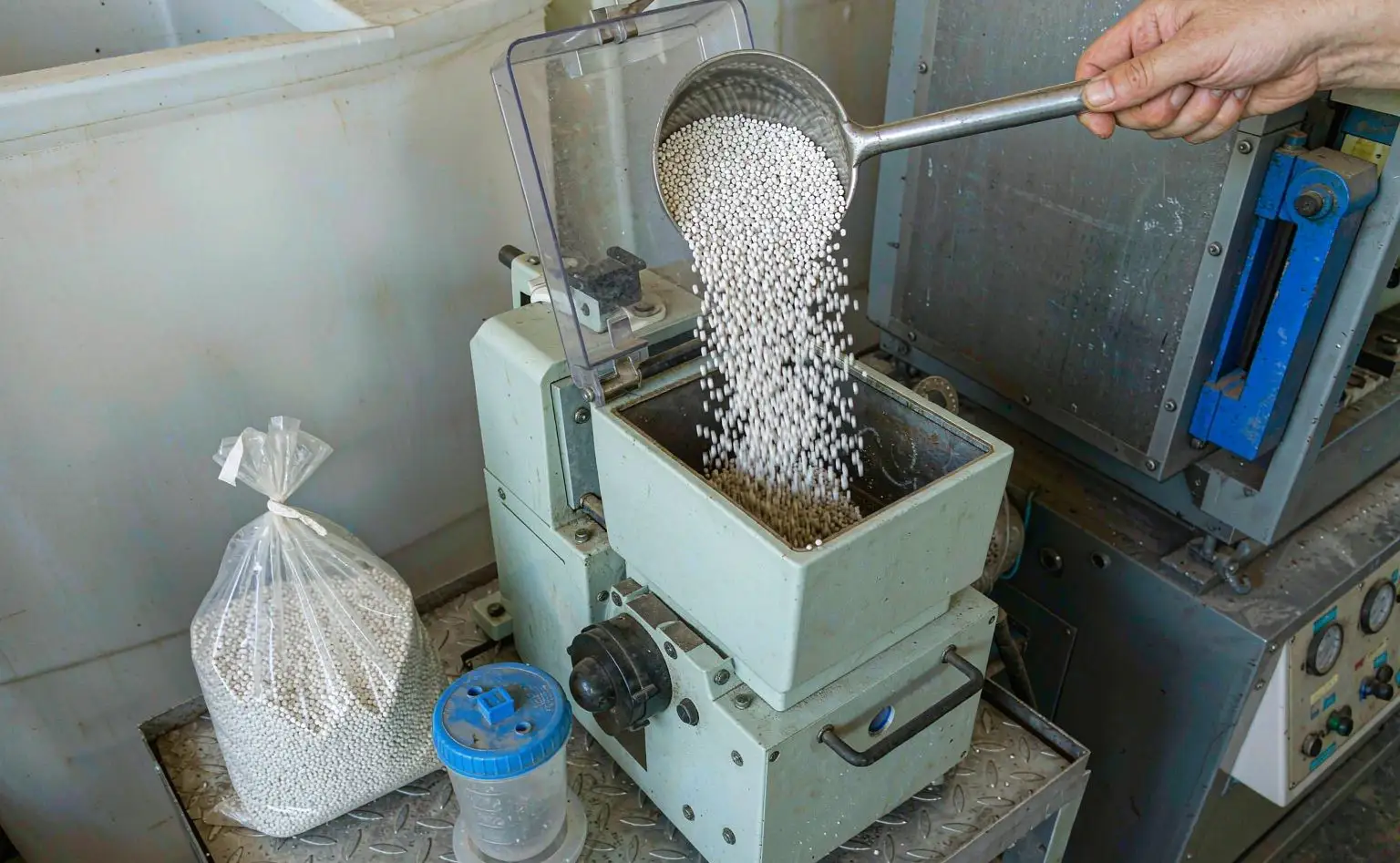 注塑机旁的操作台：工人用长柄勺把白色塑料母粒倒进注塑机料斗，旁边放着一袋开口的白色颗粒料