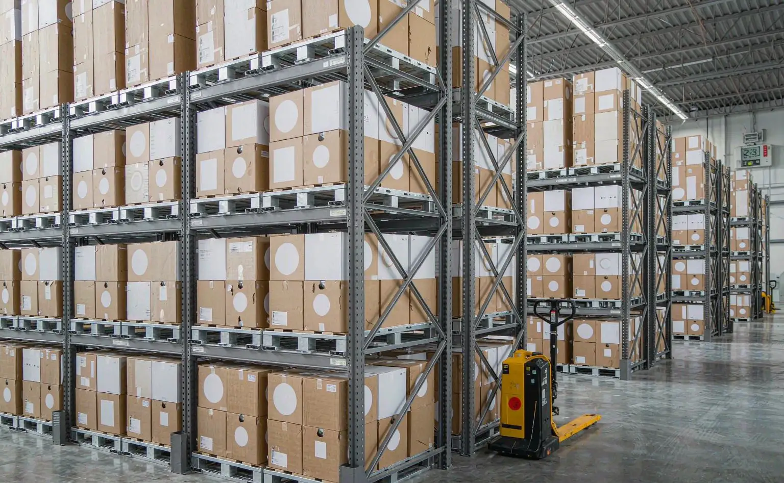 Finished-goods warehouse: palletised appliance cartons stacked on steel racking with aisle markings on the floor and an electric pallet truck parked in front