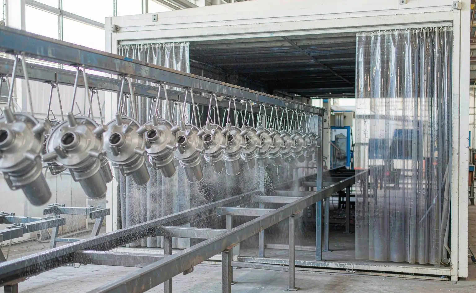 Coating shop: metal lamp holders hanging on a conveyor jig entering the spray booth, the spray covering the workpieces evenly