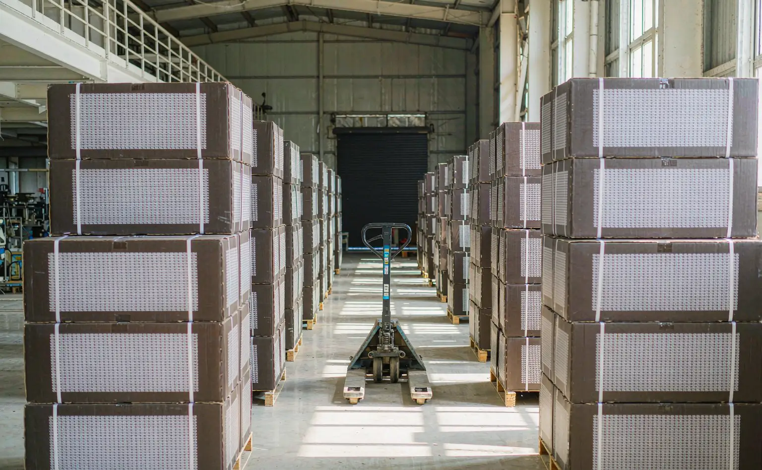 灯具成品仓库：托盘化的灯具纸箱整齐码放在车间里，箱面标签朝外，通道上停着一辆手动液压车
