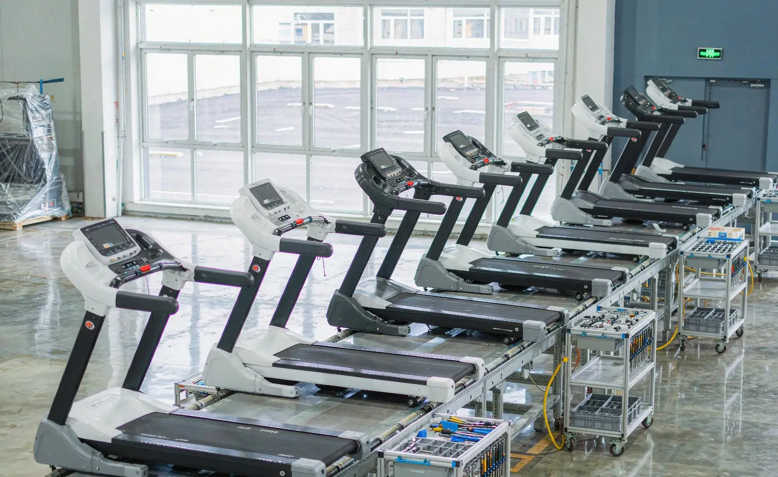 Assembly line: complete treadmills being built at the stations, the running belt already tensioned on the deck and the motor cover clipped onto the frame