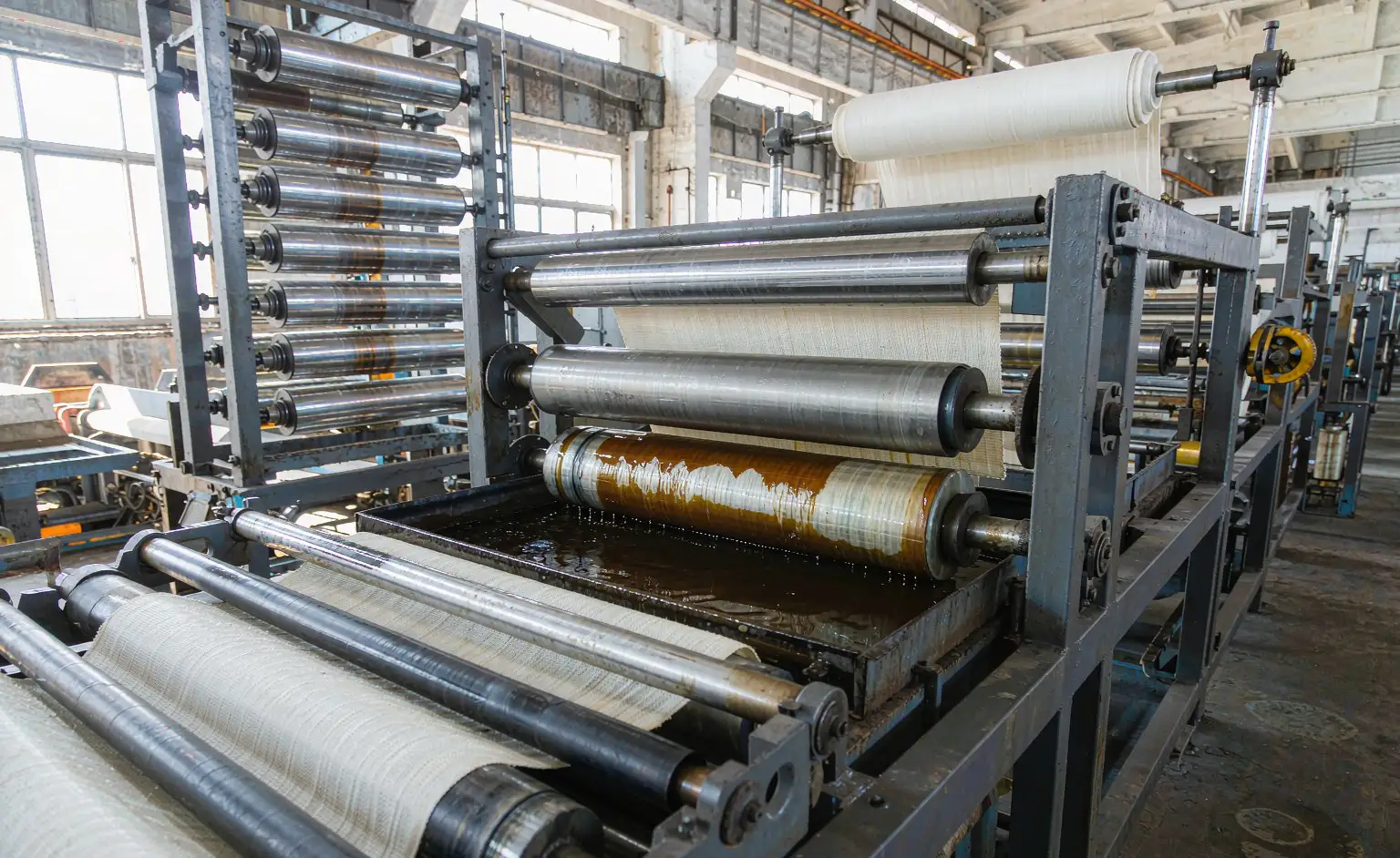 Textile shop: the running belt base fabric passing over an impregnation bath while a nip roller presses the liquor into the weave, drying cans and a winder behind it