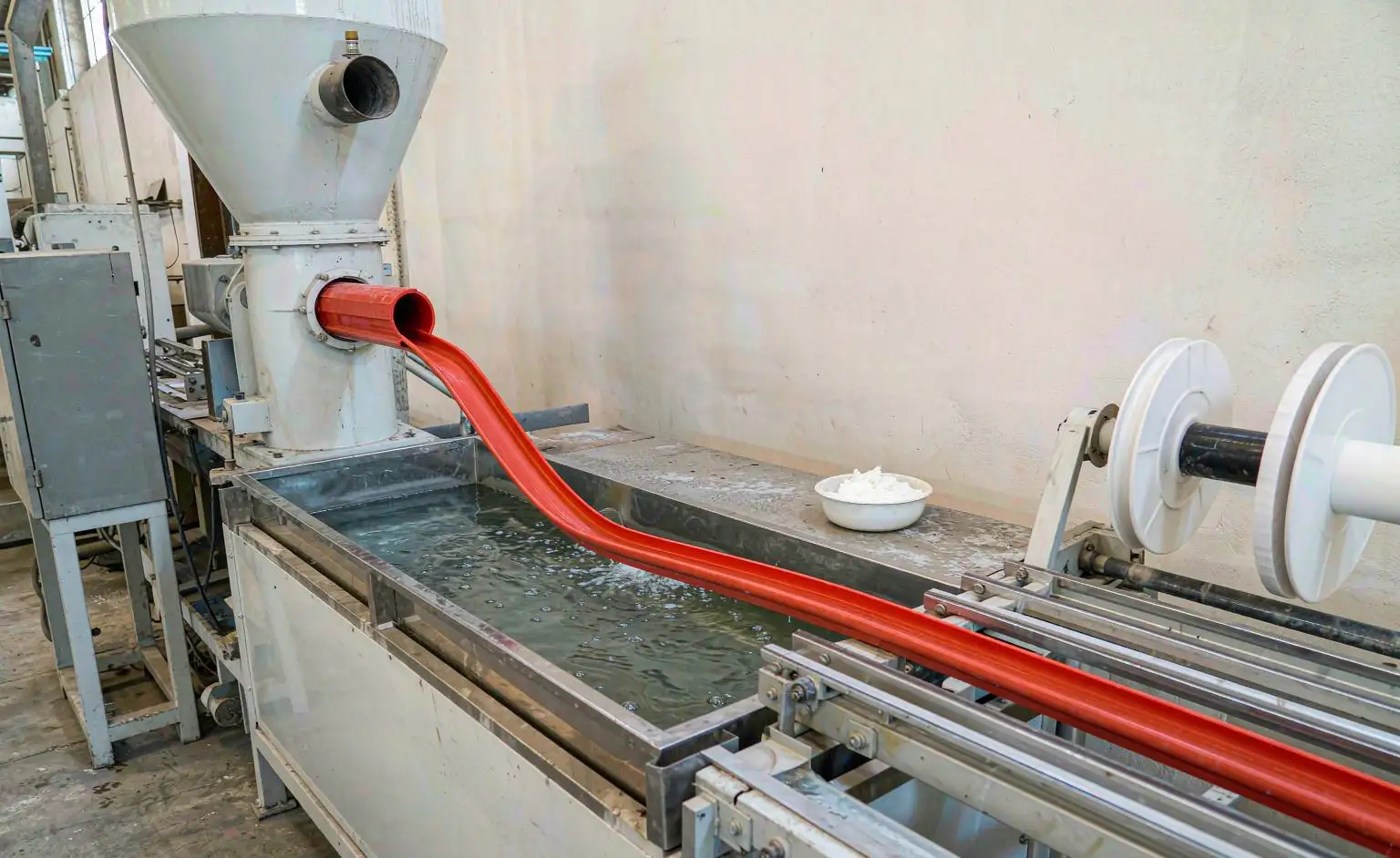 Extrusion station: PVC handrail profile coming out of the extruder die, cooled in a water bath and wound up, weighed powder additive beside the hopper