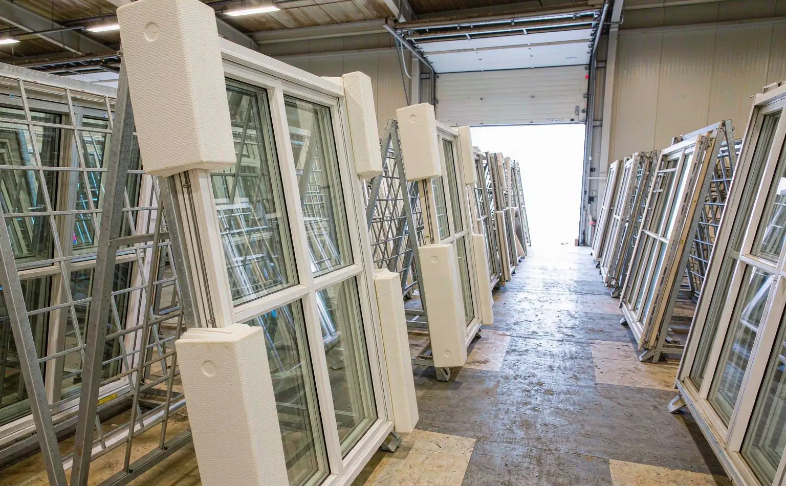 Finished-goods store: assembled aluminium doors and windows standing in steel racks, foam corner protectors between frames and aisle lines painted on the floor