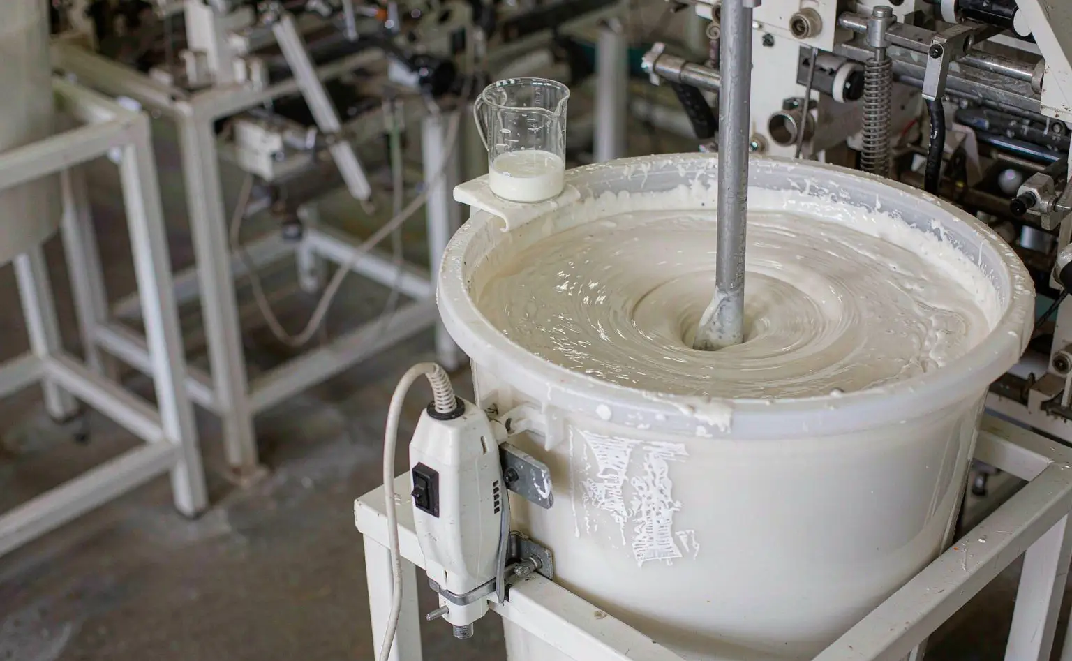 Glue station: a white plastic drum of water-based adhesive stirred into a vortex, with a measuring jug at the rim