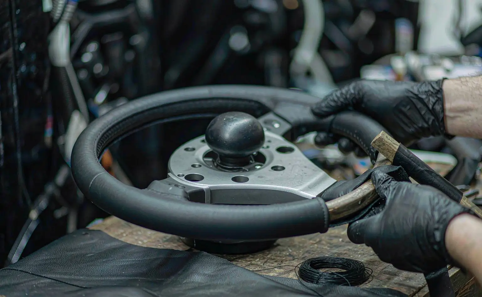 Steering wheel wrapping station: gloved hands stitching a black leather strip onto the wheel rim, thread beside it