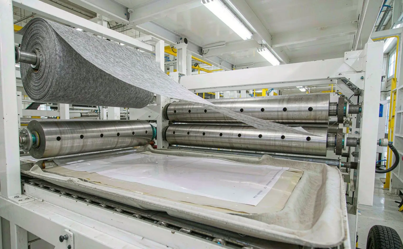 Headliner lamination line: fabric and backing feeding through hot lamination rollers, with the finished composite coiled at the output end