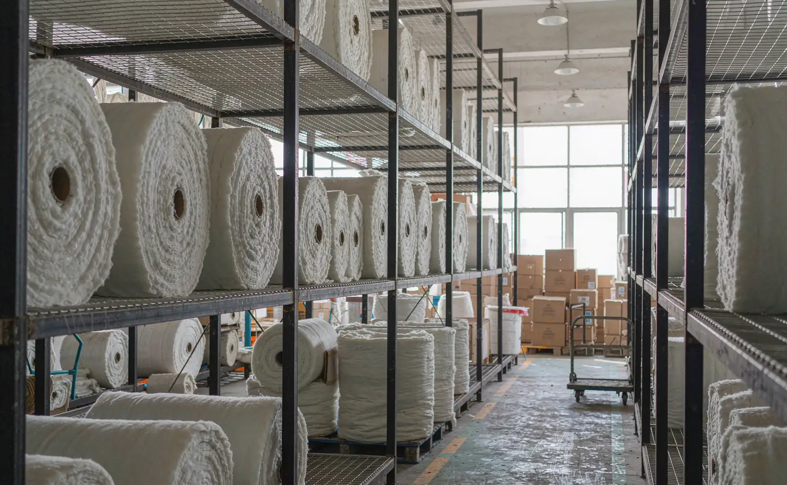 Nonwoven shop: vertical rolls of white PET cotton racked on shelves, the rolls smooth and the floor marked with aisle lines