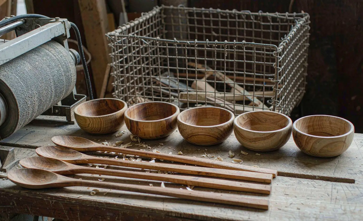 木制餐具工作台：成品木碗与木勺并排摆在木台上，碗口朝上、勺柄朝外，背景是砂光机与料筐